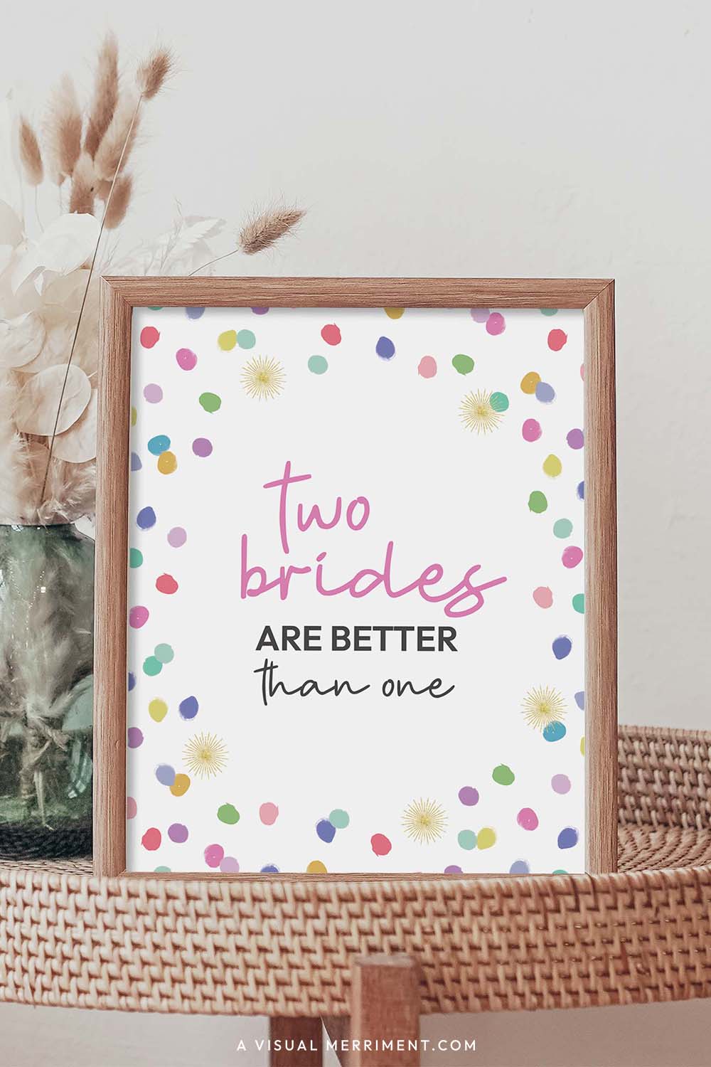 two brides better than one rainbow confetti polka dot printable party sign in a frame sitting on a grass table with a vase of boho stems