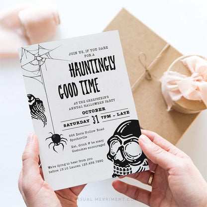 A person holds a Halloween invitation card with spooky black line art and text, with a kraft gift box and pink ribbon in the background.