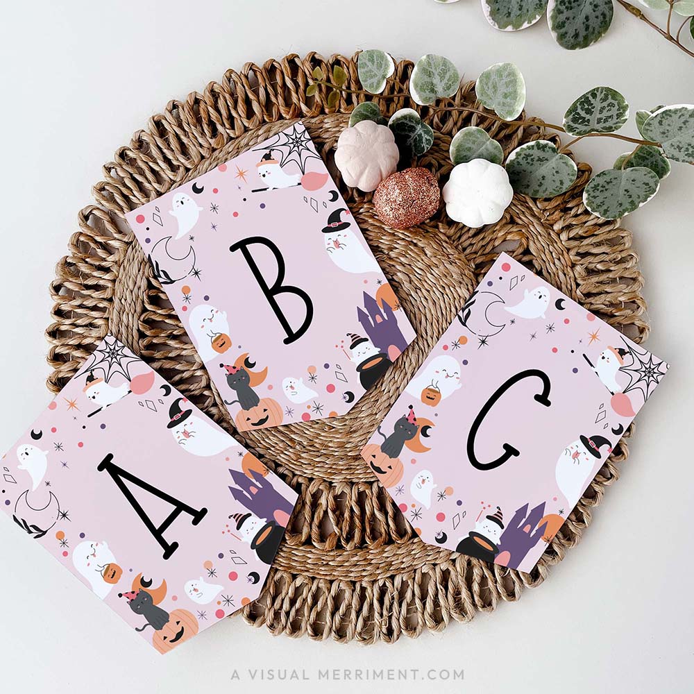 pink Halloween banner with black letters A, B, and C on flags, decorated with cute cartoon ghosts, pumpkins, witches, and spiders, arranged on round woven jute placemat