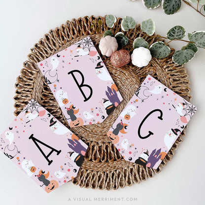 pink Halloween banner with black letters A, B, and C on flags, decorated with cute cartoon ghosts, pumpkins, witches, and spiders, arranged on round woven jute placemat