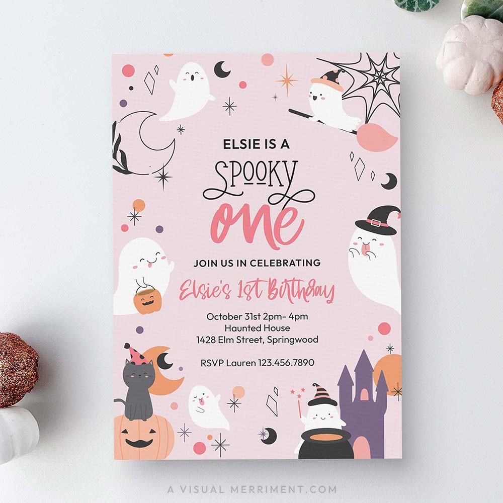 A flat-lay view of a square Halloween-themed first birthday invitation card. The card has a light pink background decorated with cute cartoon illustrations of ghosts, witches, pumpkins, a castle, and stars.