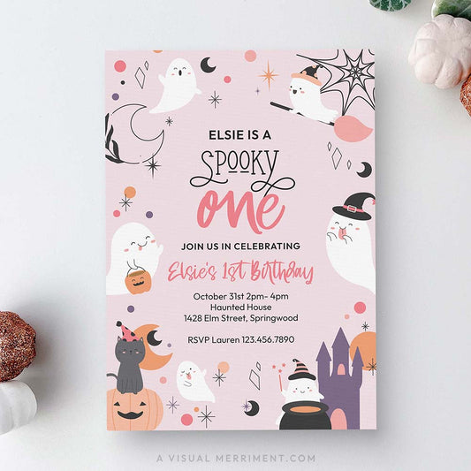 A flat-lay view of a square Halloween-themed first birthday invitation card. The card has a light pink background decorated with cute cartoon illustrations of ghosts, witches, pumpkins, a castle, and stars.