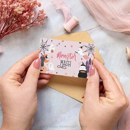 hands holding pink halloween food place card with ghost and celestial design, says monster mash, on against envelope and craft supplies