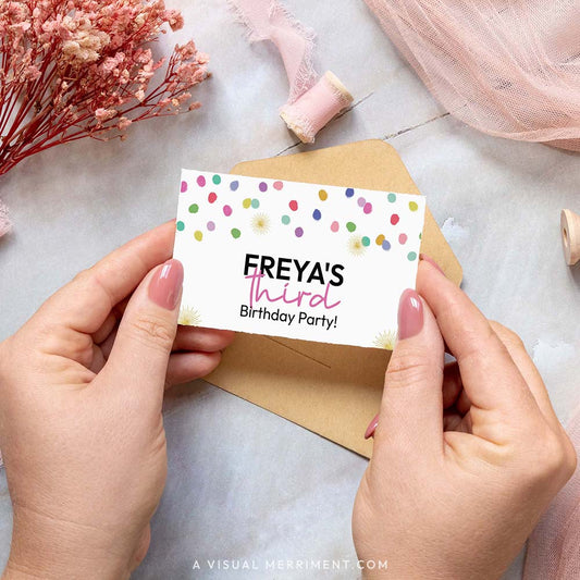 Hands holding place card with colorful polka dot rainbow confetti design, ribbons and brown envelope