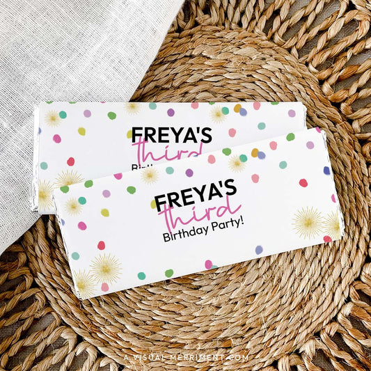 2 custom chocolate wrappers on candy bars with colorful polka dot rainbow confetti laid on a woven mat