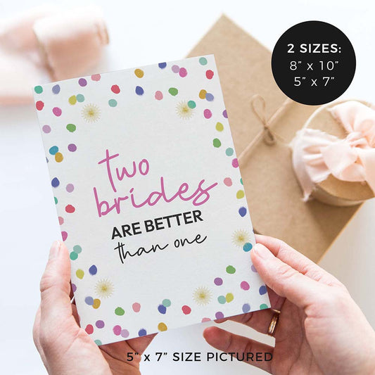 hands holding ‘two brides better than one’ wedding sign with rainbow confetti polka dot design and gifts in background