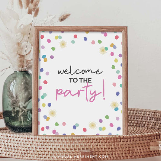 welcome to the party rainbow confetti polka dot printable party sign in a frame sitting on a grass table with a vase