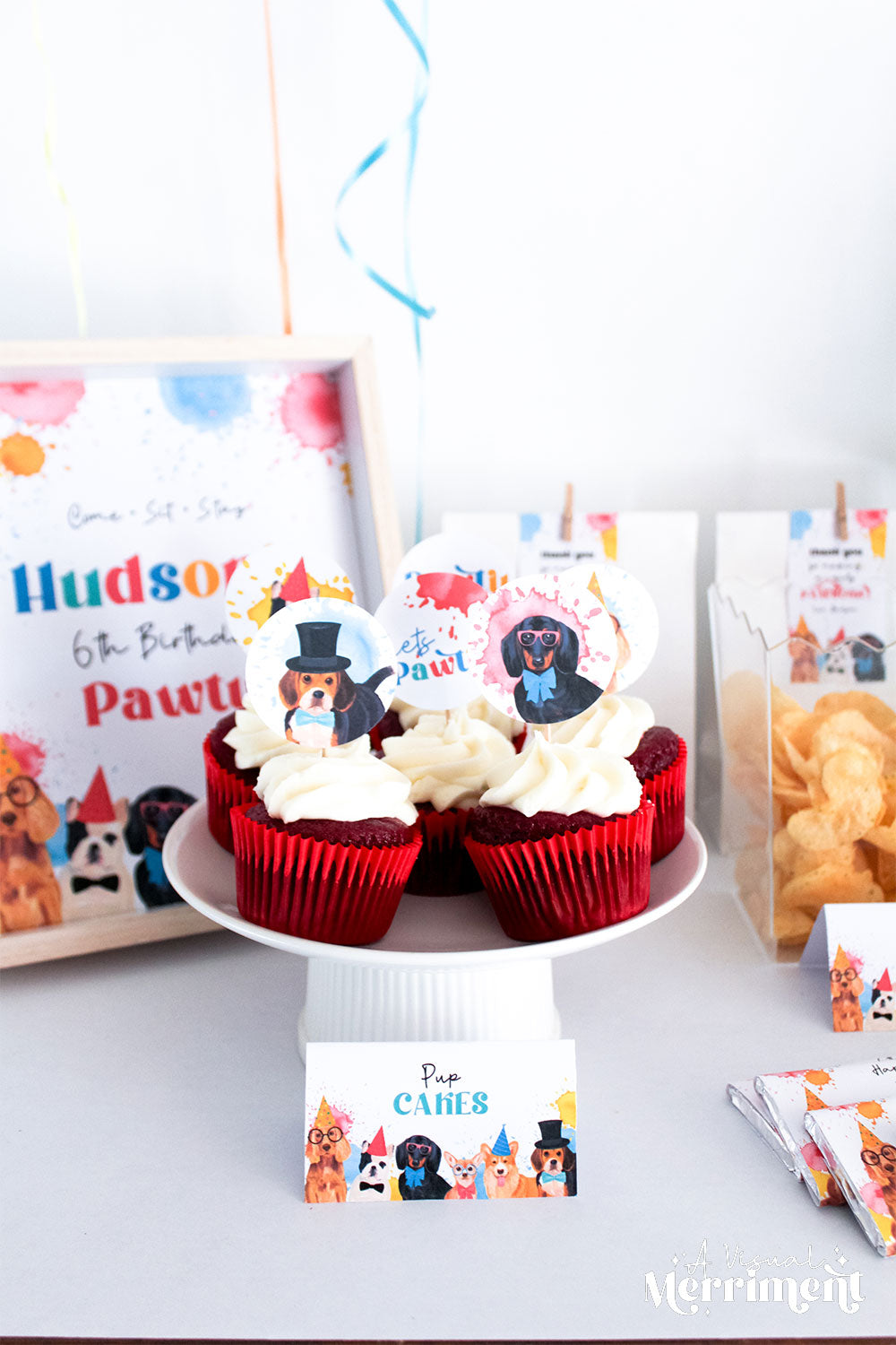 Cupcakes with dog cupcake toppers on a plate, surrounded by party supplies, signs and food
